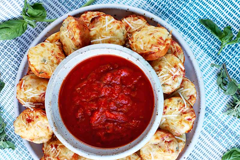 A close up top down horizontal image of a plate of mini pizza bites arranged around a bowl of tomato dipping sauce.