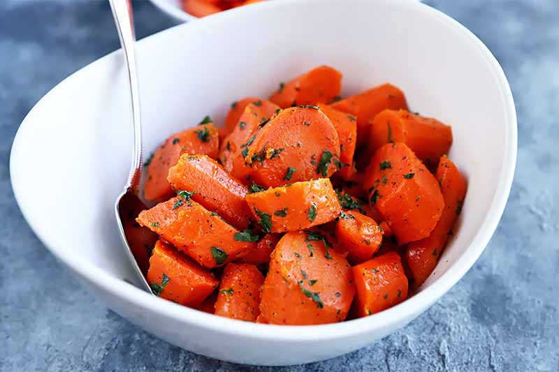 A close up horizontal image of a white bowl with freshly cooked carrots sprinkled with herbs set on a blue surface.