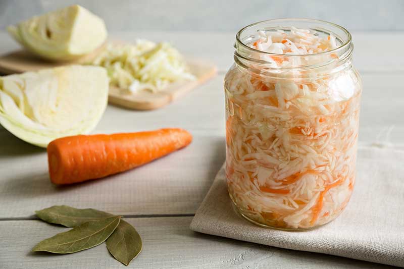 A close up horizontal image of a jar of sauerkraut with cabbage and a carrot to the left of the frame set on a wooden surface.
