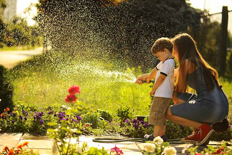 A close up horizontal image of a mother and child watering the garden with a hosepipe.
