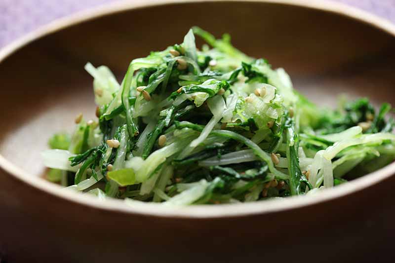 A close up horizontal image of a brown bowl filled with stir fried greens topped with pine nuts.