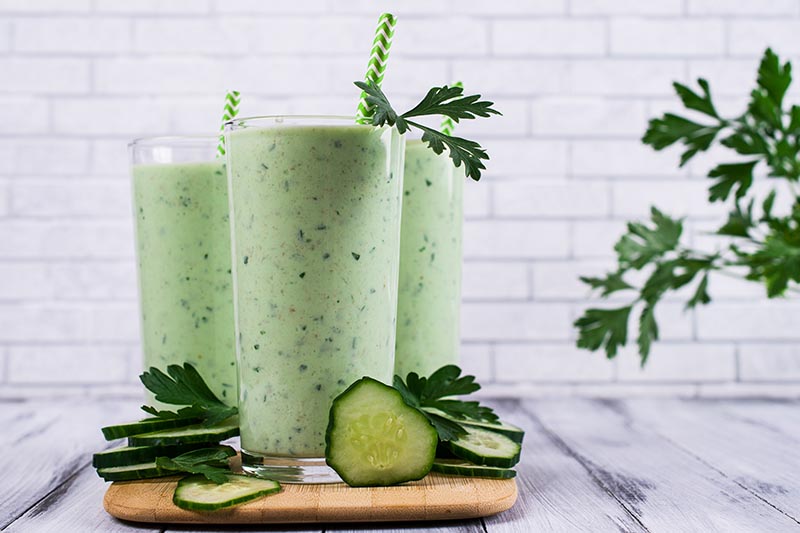 A close up horizontal image of freshly prepared cucumber smoothies set on a wooden chopping board with a white brick wall in the background.
