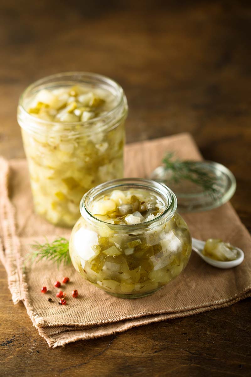 A close up vertical image of two jars of cucumber relish set on a beige fabric on a wooden surface.