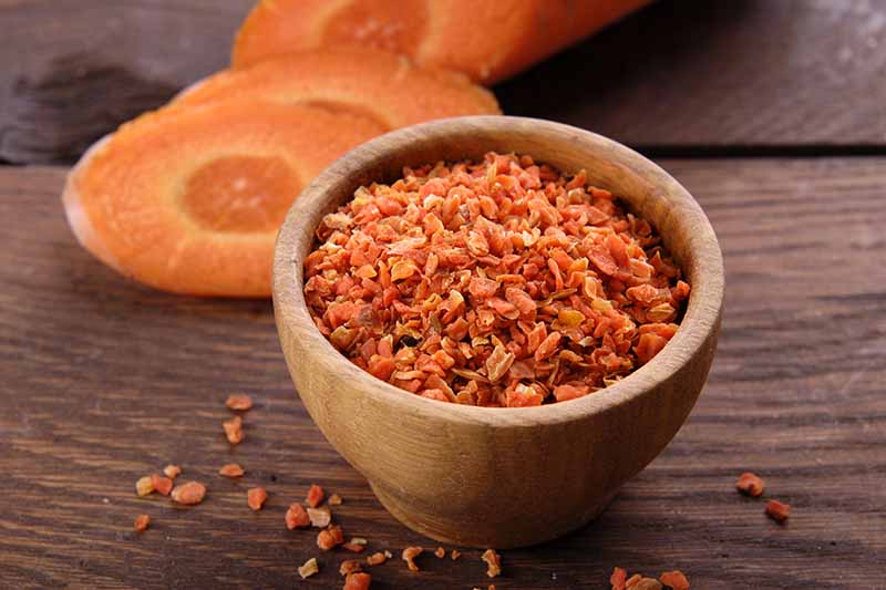 A close up horizontal image of a small wooden bowl with grated dehydrated carrot pieces set on a wooden surface.