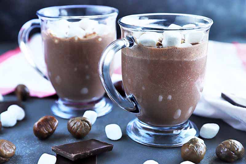 A close up horizontal image of two glass mugs filled with hot chocolate topped with marshmallows with nuts, chocolate, and marshmallows scattered around them.