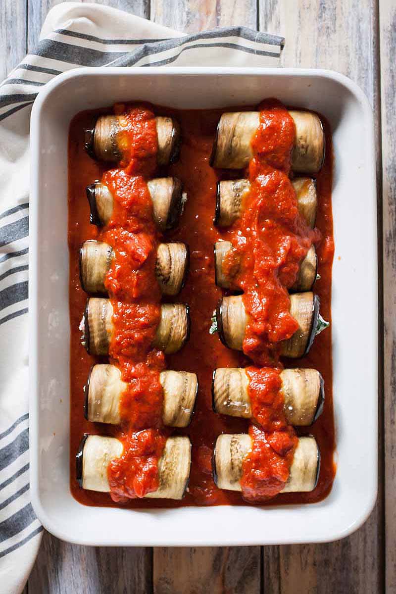 A close up vertical image of a white baking tray with homemade eggplant cannelloni set on a wooden surface.