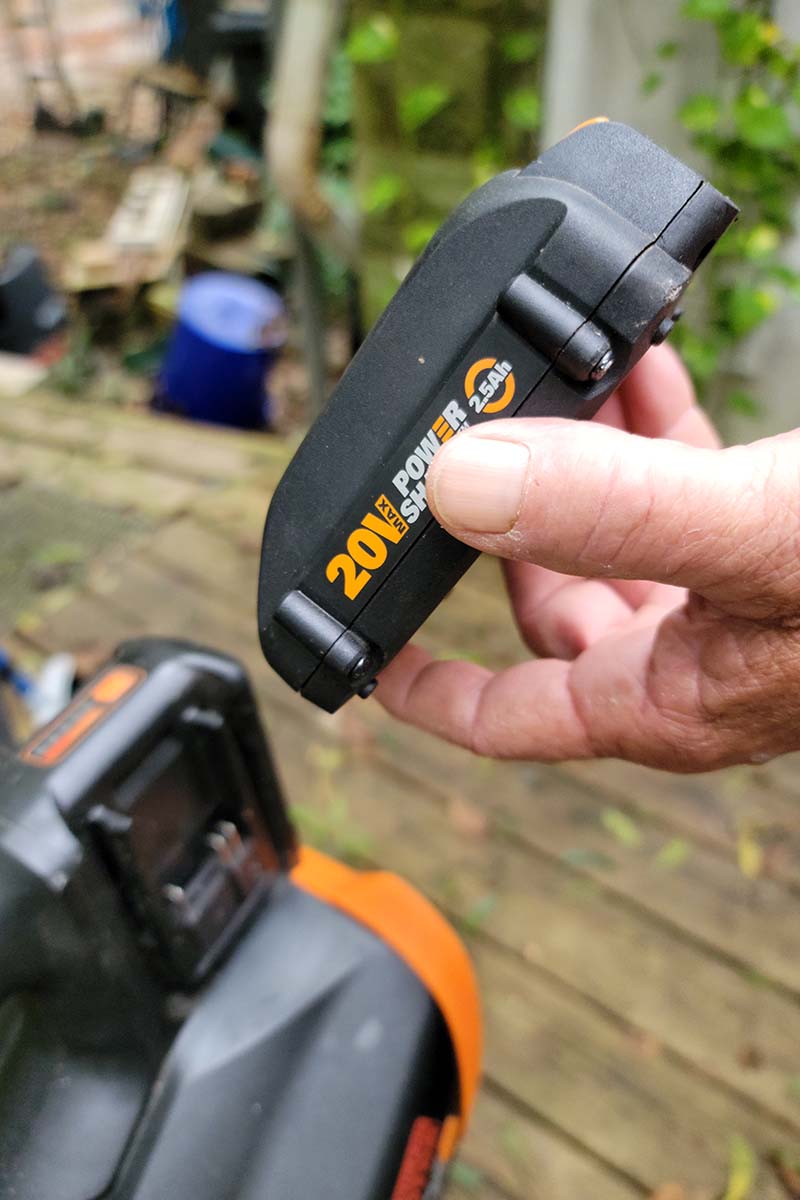 A close up vertical image of a hand from the right of the frame holding a battery to install into a leaf blower, pictured on a soft focus background.