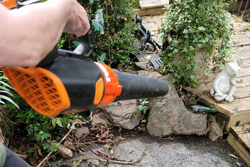 A close up horizontal image of a gardener using a cordless leaf blower to clear debris from a corner of a garden with a wooden deck in the background.