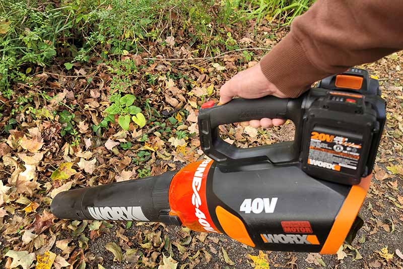 A close up horizontal image of a gardener clearing fallen leaves with a cordless electric leaf blower.