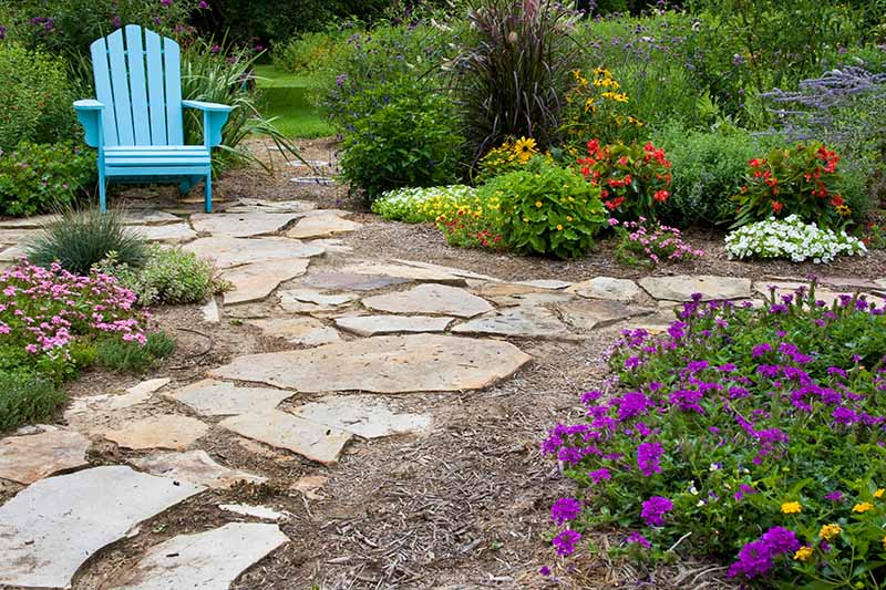 A horizontal image of a flower garden with a stone path and a blue Adirondack chair.