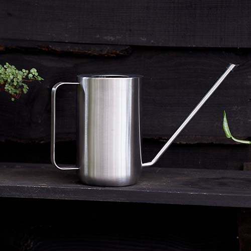 A close up square image of an elegant minimalist Blomus Modern Stainless Steel watering can set on a dark wooden surface.