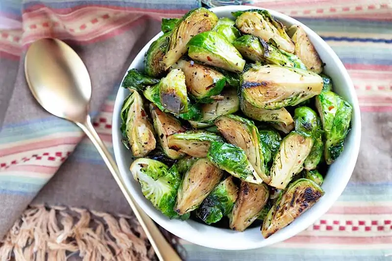 A close up horizontal image of a freshly cooked dish of caramelized red chili brussels sprouts in a white bowl with a spoon to the left of the frame.