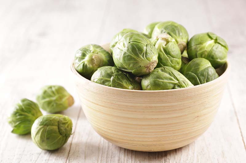 A close up horizontal image of a bowl of Brussels sprouts set on a wooden surface.