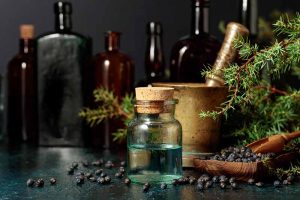 A close up horizontal image of a small glass bottle of tincture with juniper berries and branches scattered around on a dark gray table.