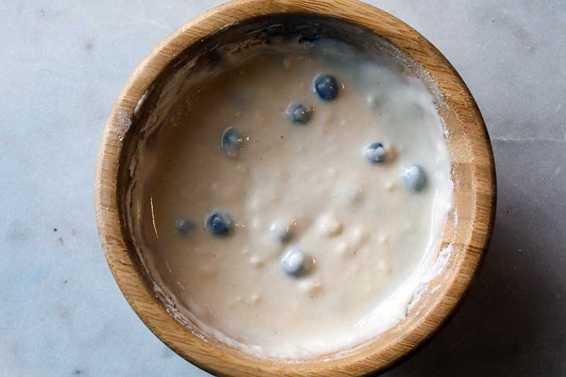 A close up horizontal image of a wooden bowl filled with juniper sourdough starter set on a gray surface.