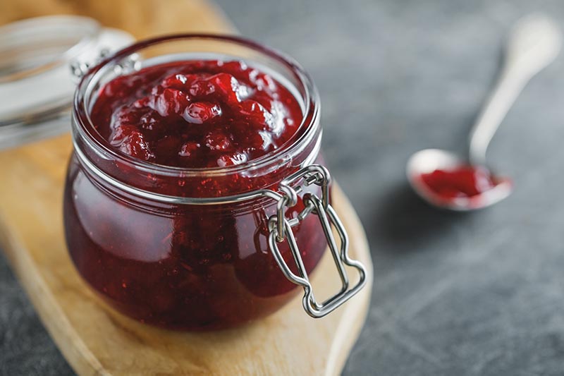 A close up horizontal image of a jar of freshly made fruit jam set on a wooden chopping board.