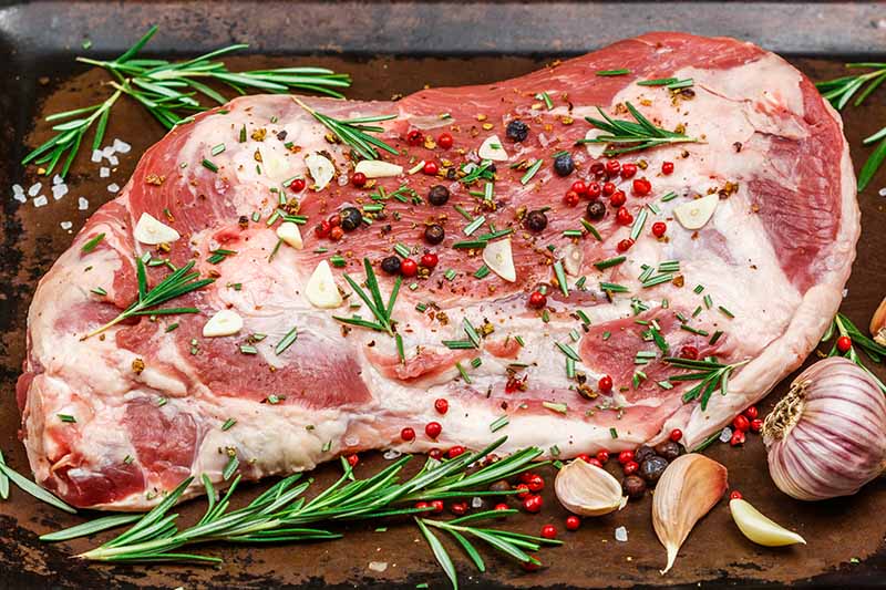 A close up horizontal image of a large leg of lamb seasoned with salt, pepper, rosemary, and juniper berries.