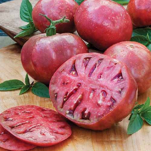 A close up square image of whole and sliced 'Cherokee Purple' tomatoes on a wooden surface.