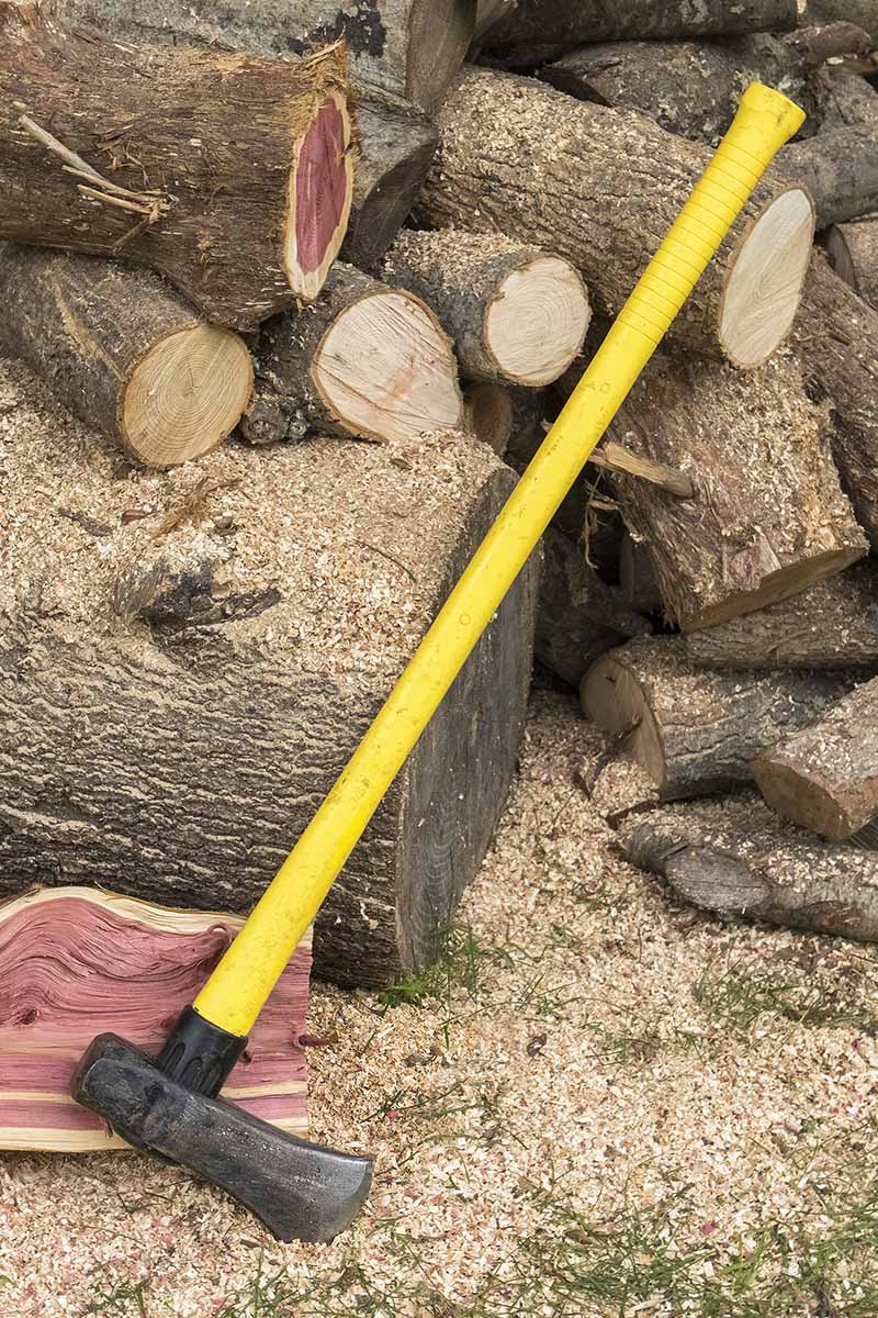 A close up vertical image of a splitting maul with a long yellow handle set on the ground with a pile of chopped wood in the background.