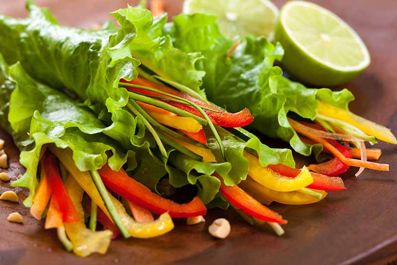 A close up horizontal image of leaf lettuce used as a wrap around a vegetables set on a wooden surface with slices of lime in the background.
