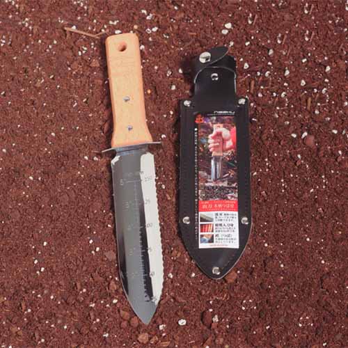 A close-up of a Nisaku Japanese garden knife set on the ground with a black leather sheath to its right.