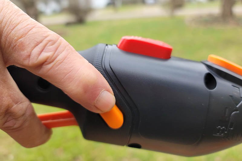 A close up horizontal image of a hand from the left of the frame holding down an orange button on the Worx WG184 to start the motor.