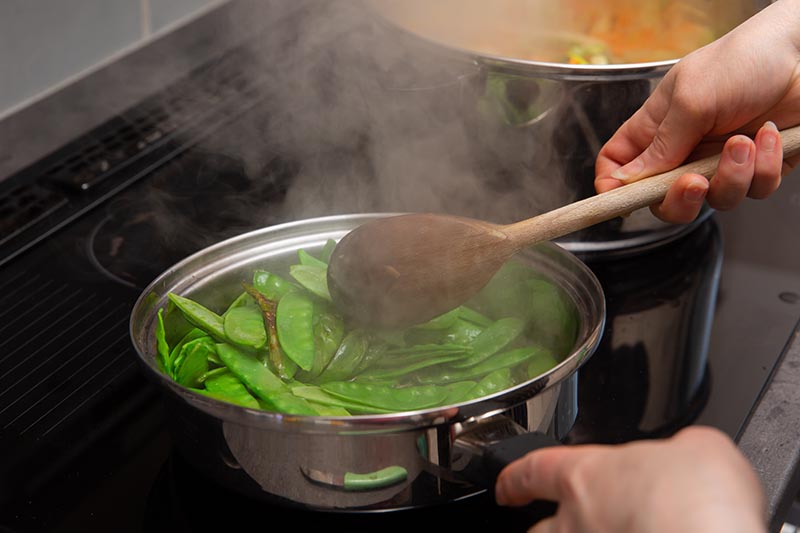 A close up horizontal image of a pot with snow peas cooking on the stove.