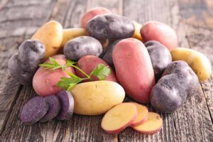 A close up horizontal image of a pile of different colored potatoes, some whole, some sliced, set on a wooden surface.
