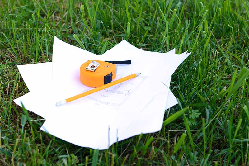 A close up horizontal image of a pile of papers with a pencil and a measuring tape set on the lawn.
