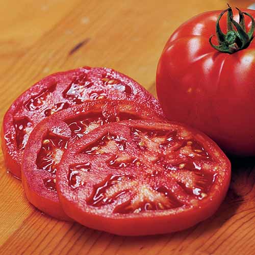 A close up square image of a whole and sliced 'Steak Sandwich' tomatoes set on a wooden surface.