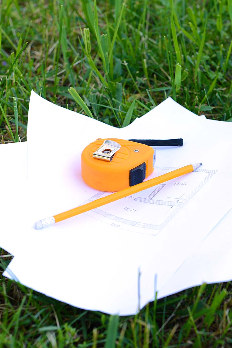 A close up vertical image of a measuring tape and paper with sketches set on the lawn.