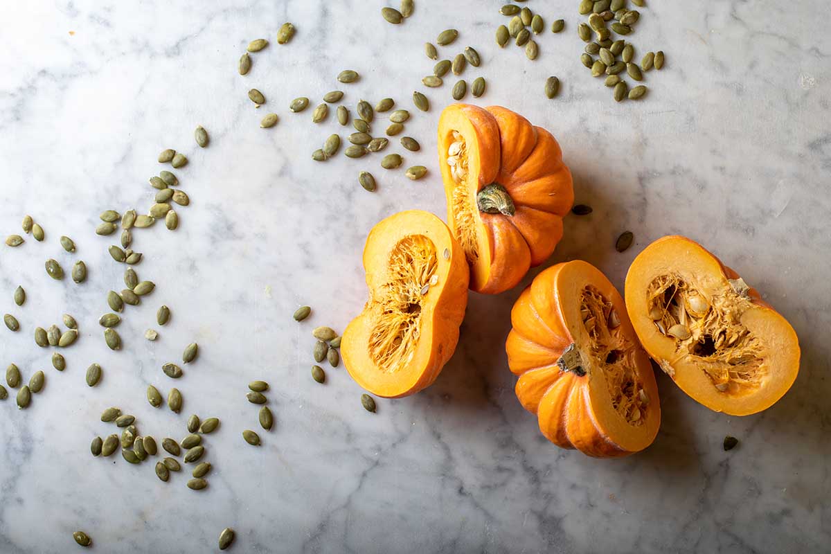 A close up horizontal image of two small pumpkins cut in half set on a marble surface with seeds scattered around.