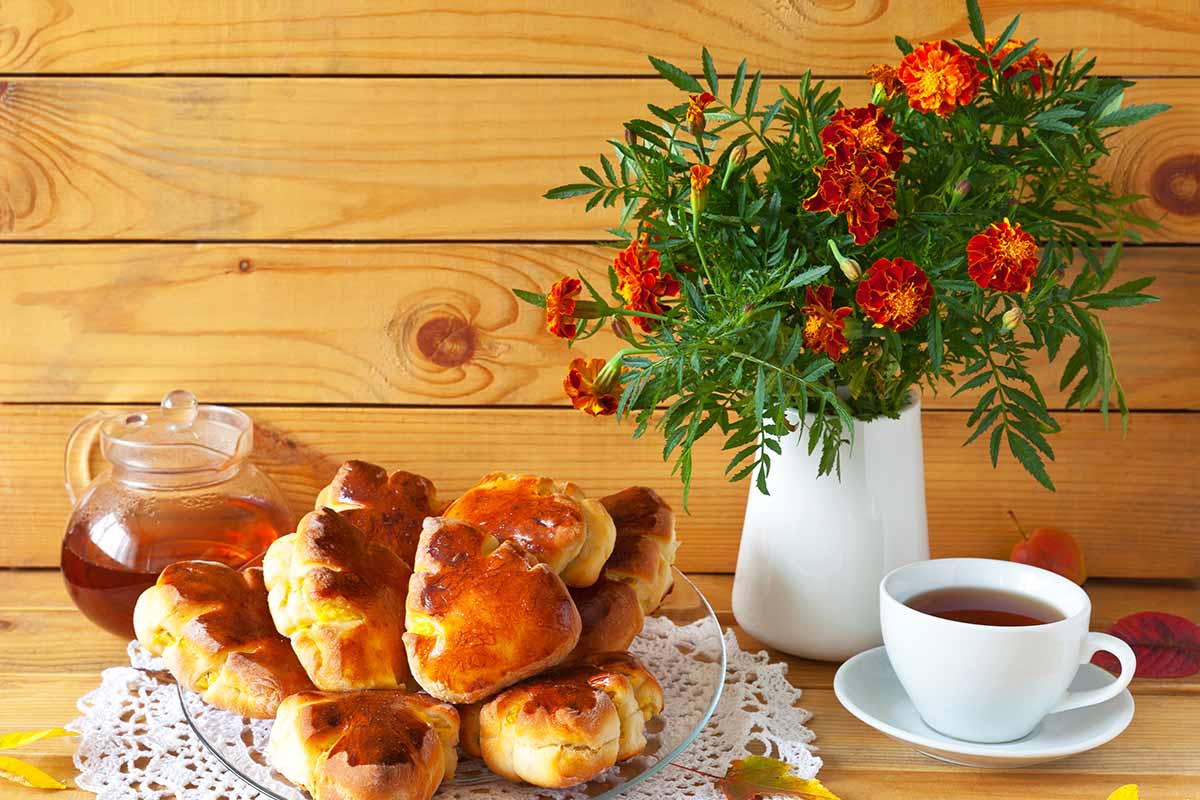 A close up horizontal image of a bouquet of marigolds with a plate of cakes and a cup of tea with a wooden wall in the background.
