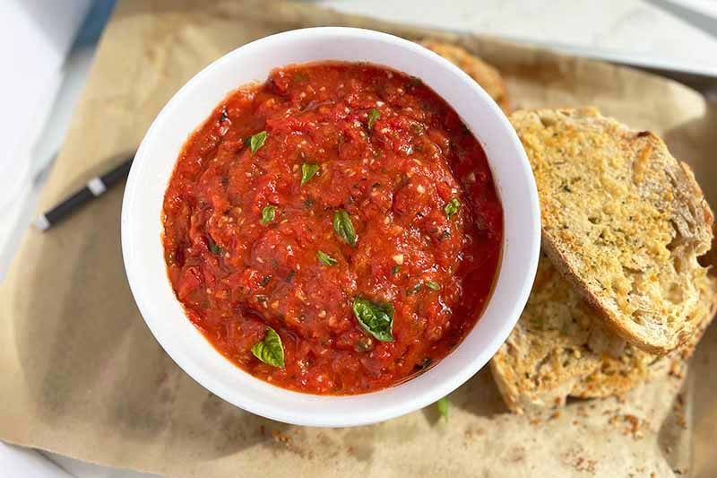 A close up horizontal image of a bowl of fresh homemade oven roasted tomato sauce with bread to the side.