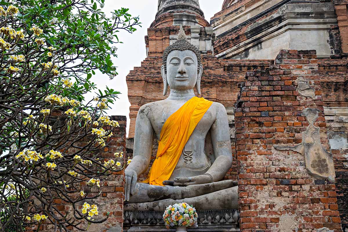 A horizontal image of a statue of Buddha outside a brick monastery with a frangipani tree to the left of the frame.