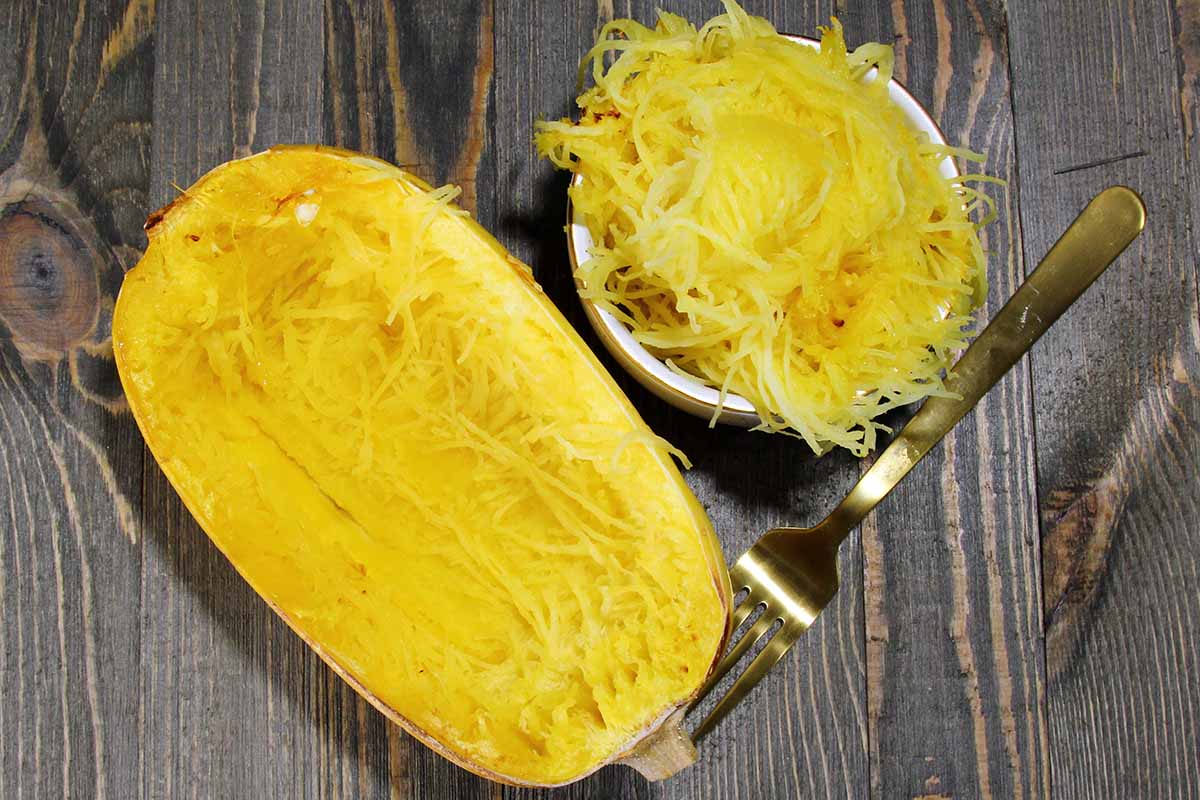 A close up horizontal image of a spaghetti squash that has been baked, cut in half, and the flesh scooped out into a white bowl.