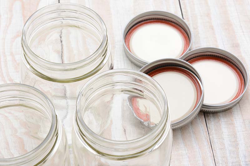 A close up horizontal image of glass canning jars and lids set on a wooden surface.