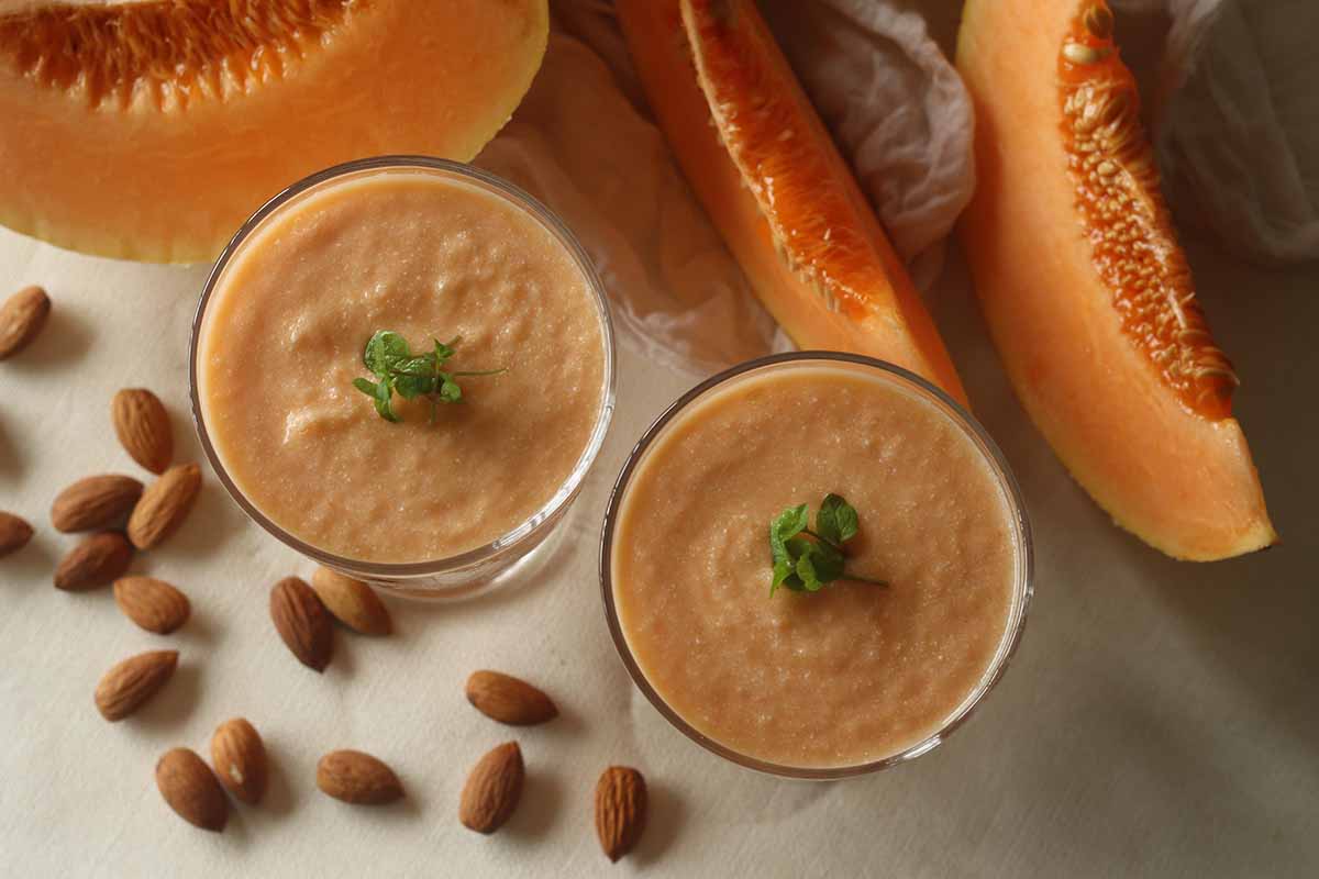A close up horizontal image of slices of melon and two glasses of freshly prepared smoothies with almonds scattered around.