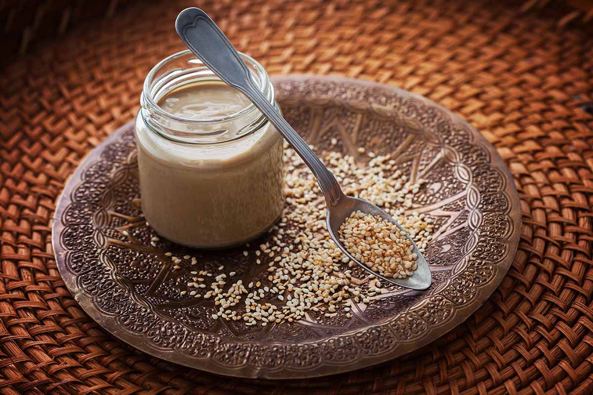 A close up horizontal image of a jar of tahini set on a decorative plate with sesame seeds scattered around.