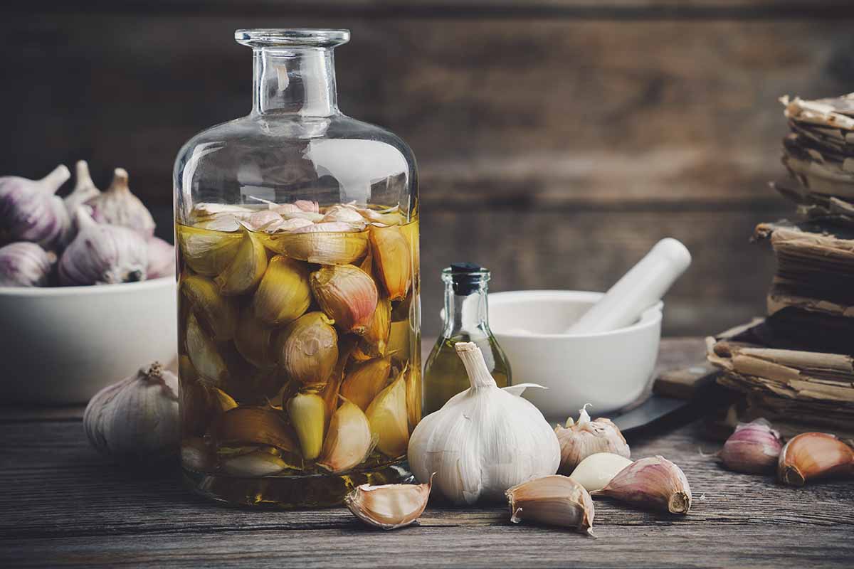 A close up horizontal image of garlic cloves in a glass jar covered with oil or water or some other liquid set on a wooden surface.