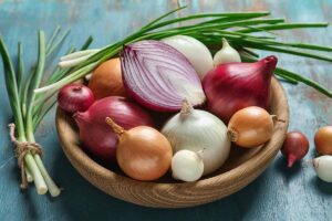 A close up horizontal image of a wooden bowl filled with different types of onions set on a rustic wooden surface.