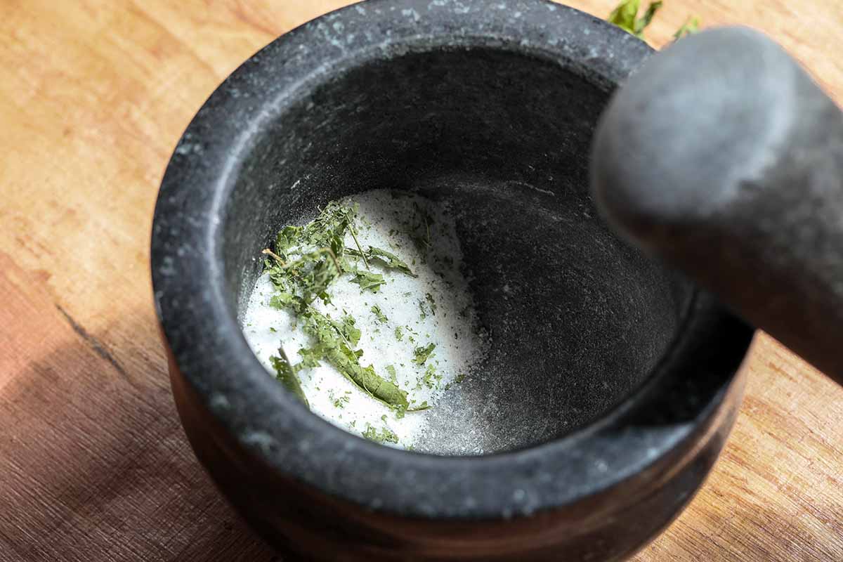 A close up horizontal image of a black pestle and mortar with herbs and sugar set on a wooden surface.
