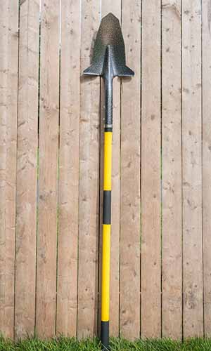 A close up vertical image of a spade leaning up against a wooden fence, rather precariously in my opinion.