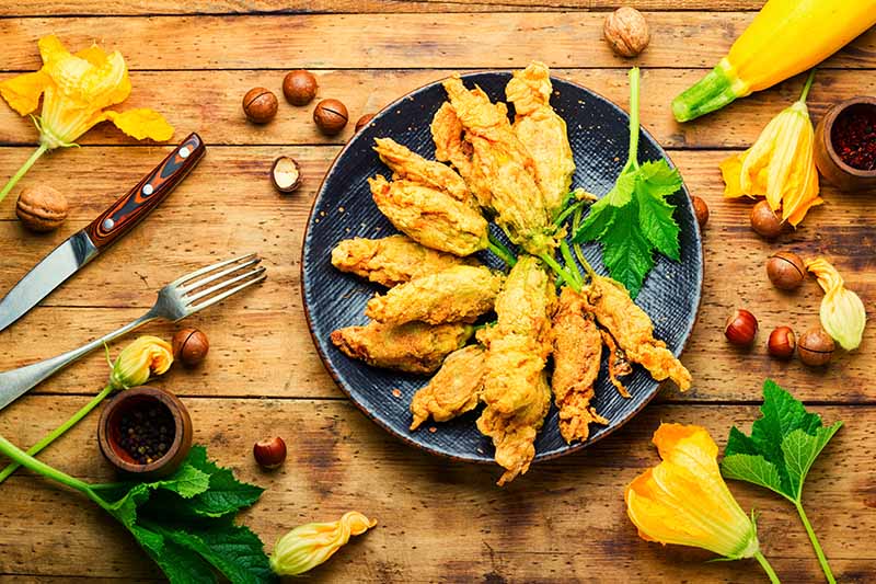 A close up of fried zucchini blossoms on a dark plate with blooms and nuts scattered around on a soft focus background.