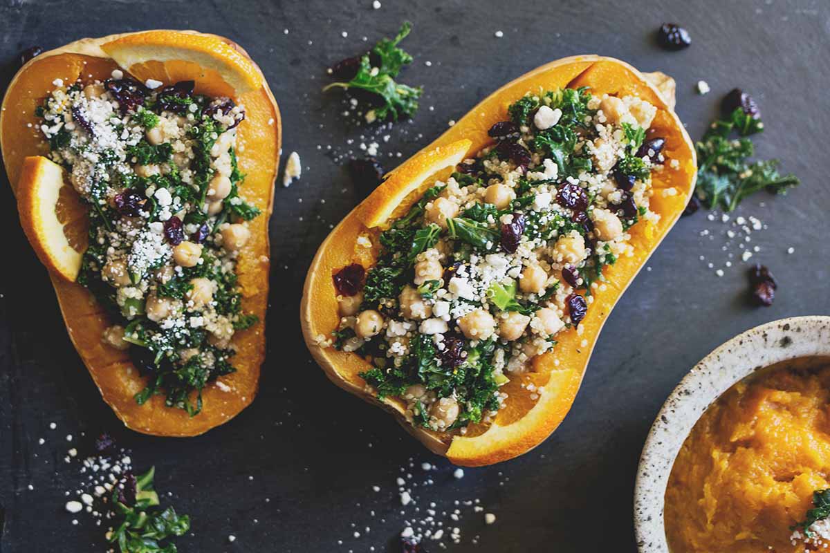 A close up horizontal image of butternut squash, halved and stuffed with quinoa, kale, cranberries, and chick peas set on a slate surface.