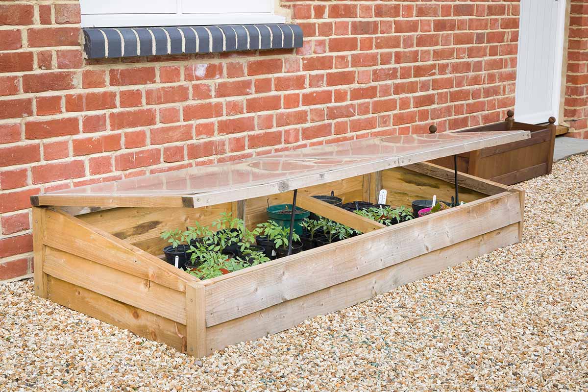 A horizontal image of a wooden cold frame propped open, set outside a brick residence on a gravel surface.