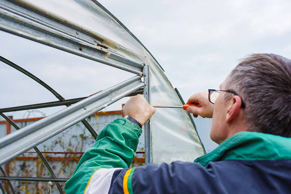 A close up horizontal image of a man repairing an old clapped out greenhouse.