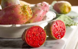 A close up horizontal image of prickly pear fruits, whole and sliced in a white bowl on a marble surface.