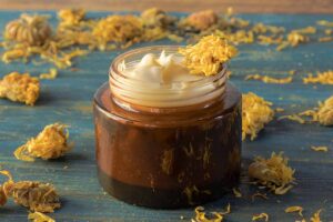 A close up horizontal image of a jar of cream with calendula flowers scattered around on a wooden surface.