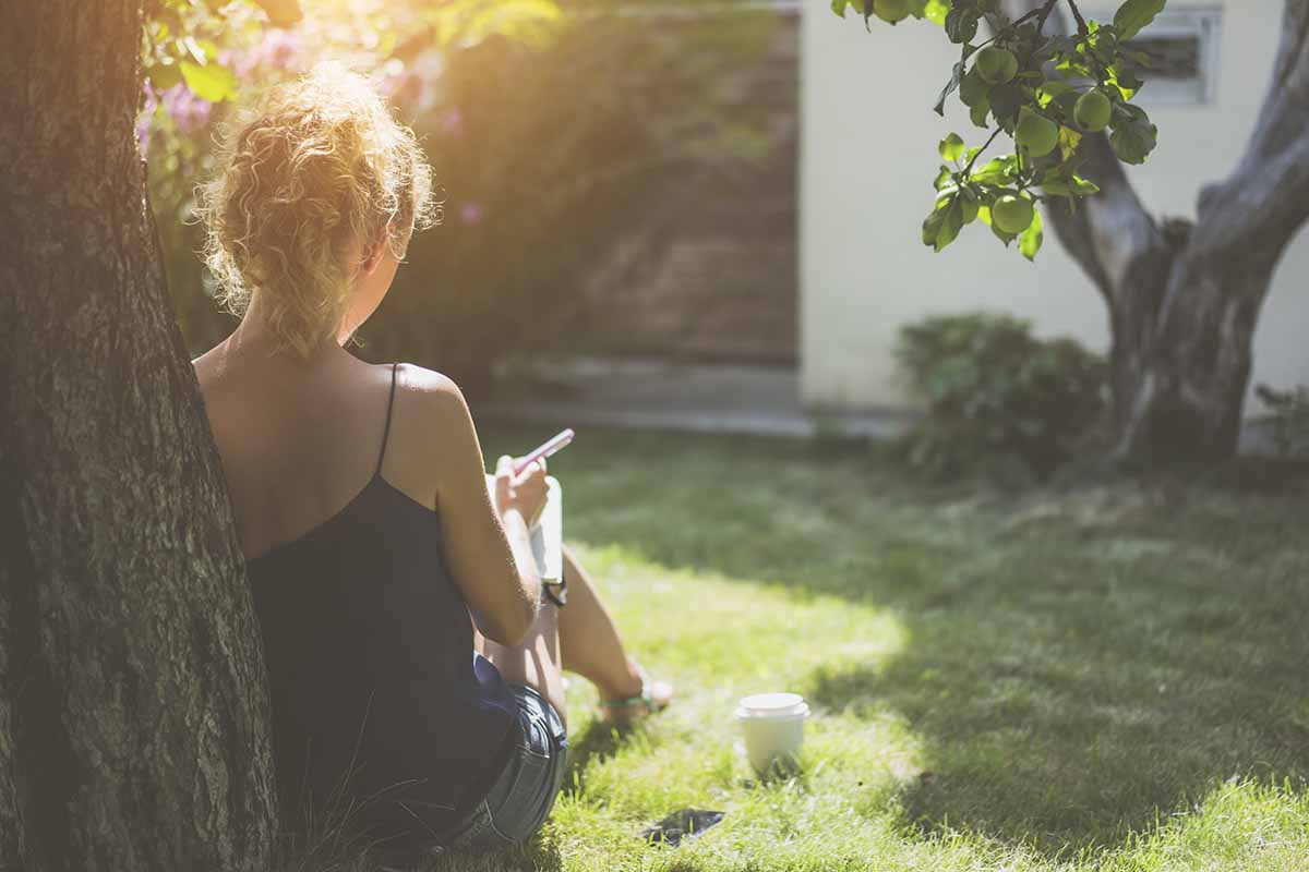 A woman, seen from the back leaning up against a tree sitting on a lawn holding a notebook and pen in light filtered sunshine with a garden in soft focus in the background.